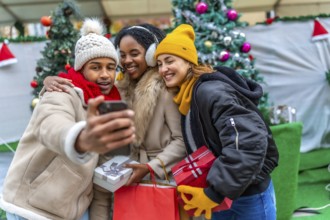 Three happy friends taking a selfie with smartphone at the christmas market, holding gifts and