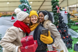 Three happy friends are taking a selfie at a christmas market, one of them is kissing another one