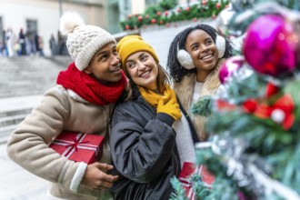 Three friends holding christmas gifts are admiring a decorated christmas tree at a city market