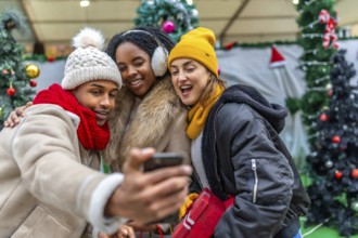 Three cheerful young friends taking a selfie with a smartphone in a city during christmas time,