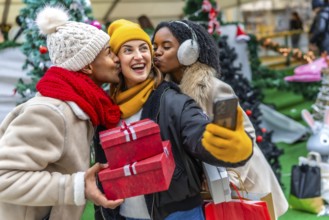 Three happy friends kissing a girl holding christmas gifts and taking a selfie at a christmas