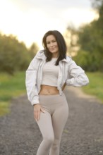 A woman stands playfully on a gravel path, wearing a light jacket, tank top and fitted leggings