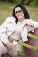 A playful woman wearing sunglasses and a light jacket is sitting comfortably on a bench in a park.