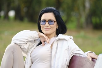 Cheerful woman enjoys a peaceful afternoon in a park, wearing sunglasses and a light jacket. She