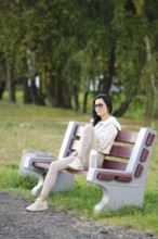 A slender woman sits comfortably on a park bench, wearing glasses and casual attire. Sunlight