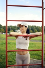 A woman leans against red workout bars in a park, enjoying the sunlight. She wears activewear and a