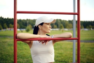 A woman in a casual outfit stands at a fitness station in a park, resting her arms on the bars