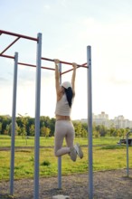 A woman is performing an exercise on pull-up bars in a city park. The sun is setting, casting a