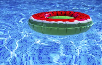 A bright ring-a float, with a watermelon, in the clear blue water of the pool, under the sunlight