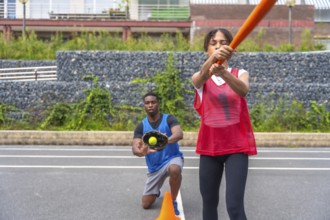 Engaging in an outdoor baseball practice, a woman swings a bat with focus while a man kneels,