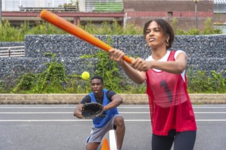 Female baseball player swinging a bat to hit a ball while a male catcher prepares to catch it.