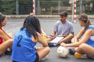 Soccer coach sitting on the ground, using a notebook to explain game strategies to female athletes