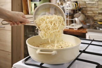 A hand adds cooked noodles from strainer in a pot of creamy mushroom sauce on the stove