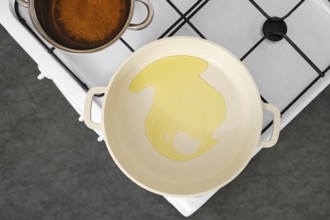 Cooking oil is poured into a frying pan on a stovetop. A pot with vegetable stock sits nearby,