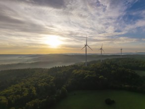 Wind turbines rise above a misty forest at sunrise, a symbol of renewable energy in a peaceful