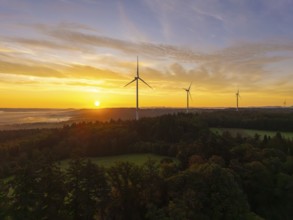Wind turbines in a hilly and wooded landscape, illuminated by the rising sun, near Schorndorf,