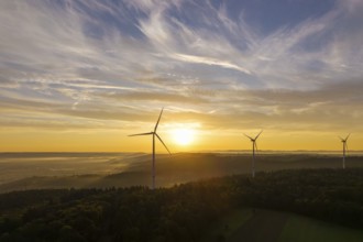 Wind turbines stand still under a soft sunlight illuminating the sky and forest, near Schorndorf,