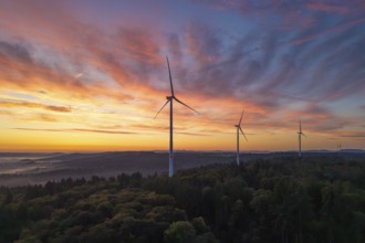 Three wind turbines in the foreground of a colourful morning sky over a hilly landscape, near