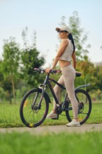 A woman stands with her bicycle in a green park, wearing athletic clothing and sunglasses. She