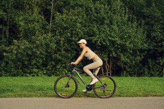 A woman rides a bicycle along a smooth path with vibrant green grass on one side and dense trees