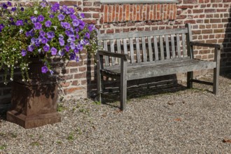 A wooden bench stands next to a lush flowerpot with purple flowers in front of a brick wall in the