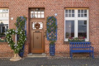 A decorated brick house façade with a blue bench and floral decorations next to the entrance, North