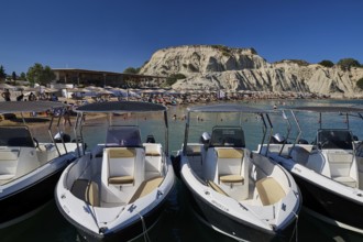 Several boats are moored on a jetty with a rocky beach in the background. Holidaymakers relaxing on