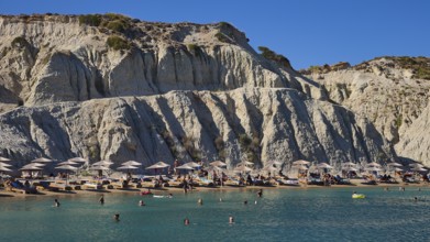 Beach in front of a large rock face, lined with sun loungers and umbrellas, bathers enjoying the
