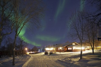 An illuminated house under auroras with snow-covered trees on a winter night, Sortland campsite,