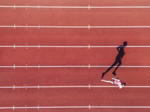 Aerial view of a female individual (runner) on a red athletics track (tartan track) with shadow as