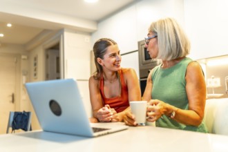 Mother and daughter chatting and smiling while enjoying coffee and browsing a laptop together in a