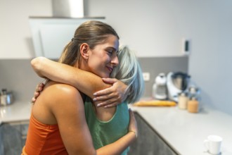 Happy adult daughter is embracing her senior mother in a kitchen, showing love, care, and support