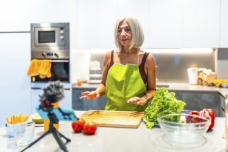 Mature woman in a green apron records a cooking tutorial in a modern kitchen, surrounded by fresh