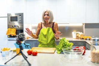 Elderly woman in a green apron recording a cooking tutorial with fresh vegetables, showcasing