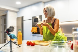 Elderly woman wearing an apron preparing healthy food in her modern kitchen and recording a video