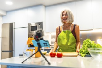 Smiling mature woman wearing apron preparing recipe for her cooking vlog, recording video with