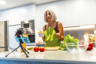 Cheerful mature woman wearing apron holding bowl of carrot sticks while recording video blog about