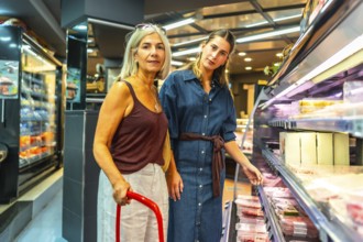 Two women shopping for groceries are choosing meat products from refrigerated section of a