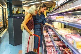 Two women selecting fresh products from a refrigerated section in a grocery store, using a basket