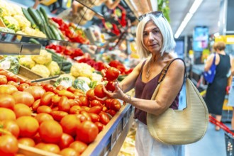 Mature woman choosing ripe tomatoes in a vibrant grocery store produce aisle, surrounded by an