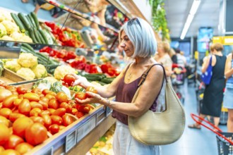 Mature woman with gray hair choosing ripe tomatoes in a grocery store, surrounded by colorful
