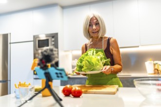 Senior woman creating a cooking video, holding fresh lettuce and surrounded by vegetables in a