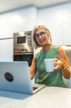 Smiling senior woman enjoying coffee while working on her laptop in a cozy kitchen, embracing a