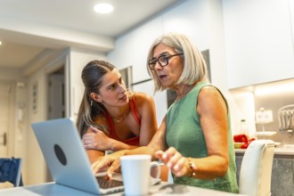 Young woman assisting her senior mother with a laptop in a home kitchen setting, providing