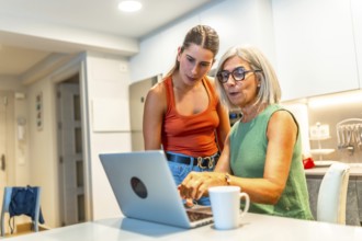 Senior woman navigating a laptop with help from a younger woman in a cozy kitchen, fostering family