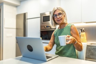 Smiling senior freelancer woman enjoying a cup of coffee while working on her laptop in her modern