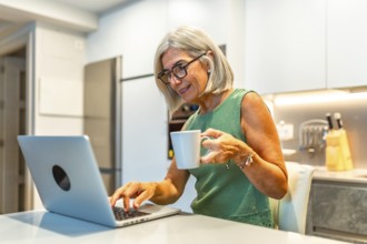 Smiling senior woman enjoying coffee while working on a laptop, embracing the comfort of her home