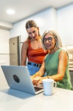 Senior woman using a laptop with guidance from a young woman in a modern kitchen, sharing knowledge
