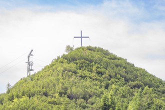 A cross is enthroned on a green mountain peak under a slightly cloudy sky, Herzogstand summit,