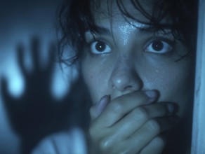 A frightened woman with a shadowy figure in the background holds her hand in front of her mouth,